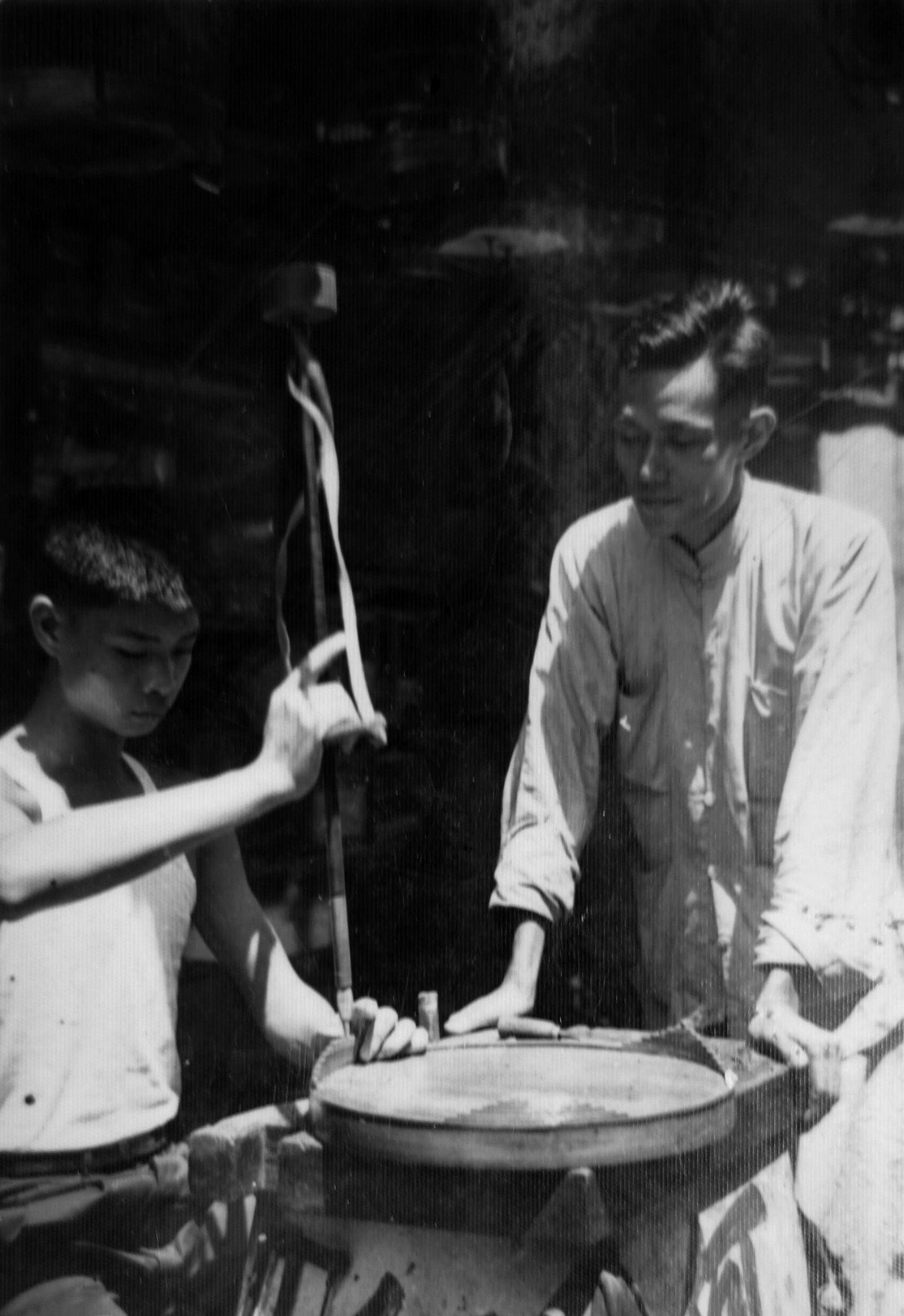卓康師傅(右)正在指導手持鑽孔工具的徒弟陳樂財(左)製作鳥籠,約攝於1950年代末。